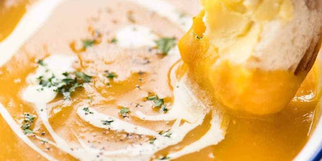 Close up of dunking crusty bread into thick and creamy pumpkin soup in a rustic white enamel bowl.