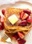Pile of French Toast on a rustic white plate, doused with maple syrup and topped with a pat of melting butter and strawberries.