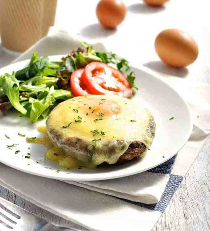 Baked Eggs in Mushrooms covered in melted cheese on a white plate with coffee on the side, ready to be eaten.