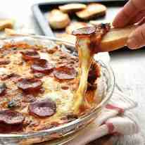 Toasted bread scooping up ultra cheesy, gooey Pizza Dip in a glass dish with a great cheese pull shot.