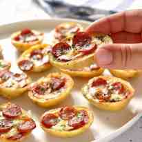 Close up of a hand holding a Mini Pizza Potato Skin with a platter of Mini Pizza Potato Skins in the background.
