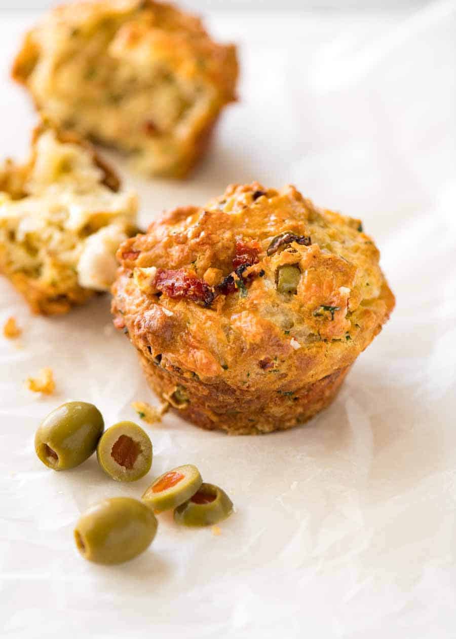Savoury Cheese Muffins with Sun Dried Tomatoes, Feta and Olives on white paper, with olives on the side, ready to be eaten.