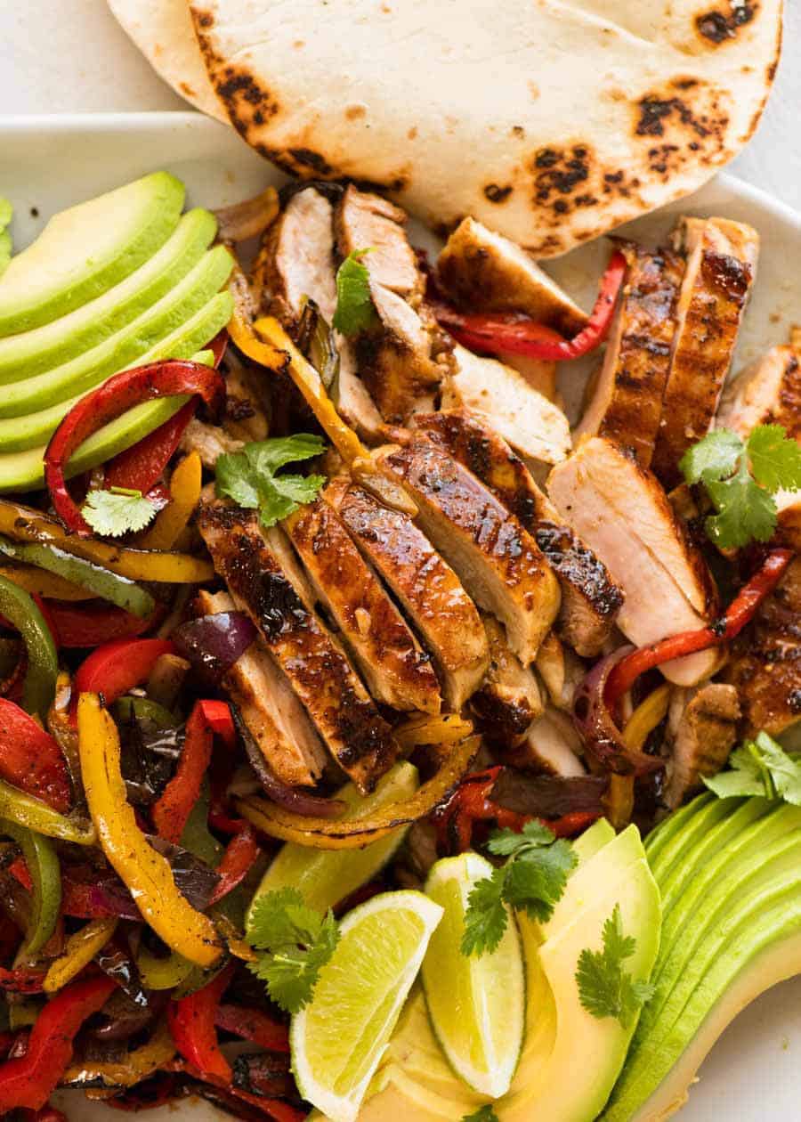 Close up of sliced Chicken Fajitas alongside charred peppers, avocado and warm tortillas, ready to be served.