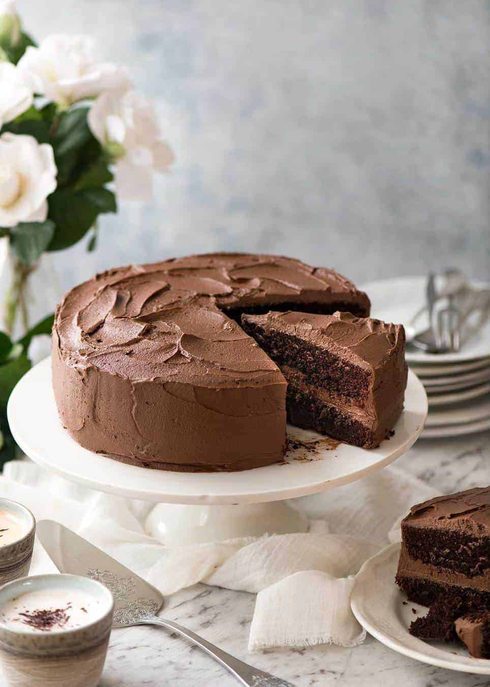 A moist Chocolate Cake with Chocolate Buttercream Frosting on a white cake platter with a slice cut and partially pulled out and a stack of dessert plates next to it.