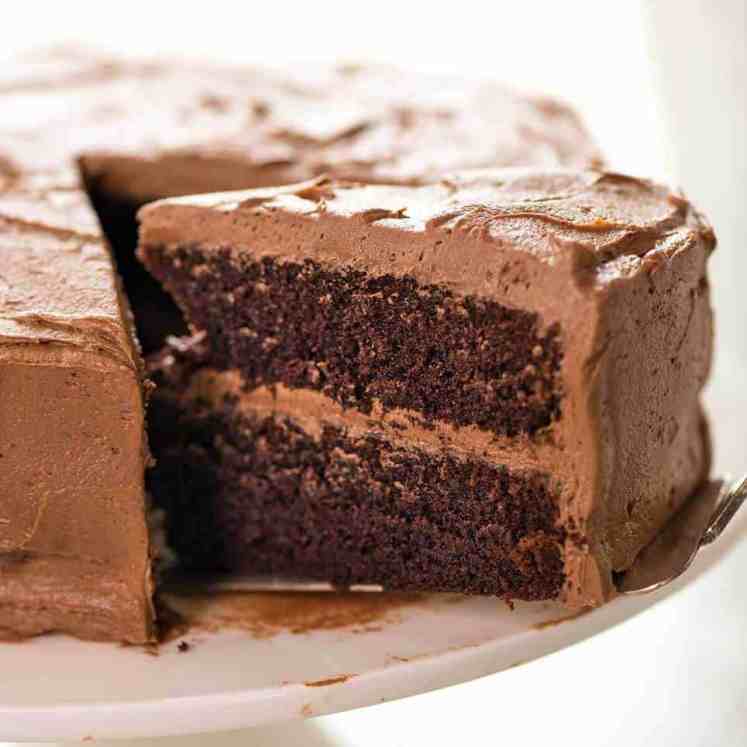 A photo showing half a Chocolate Cake frosted with Chocolate Buttercream on a white cake stand with a large slice partially pulled out.