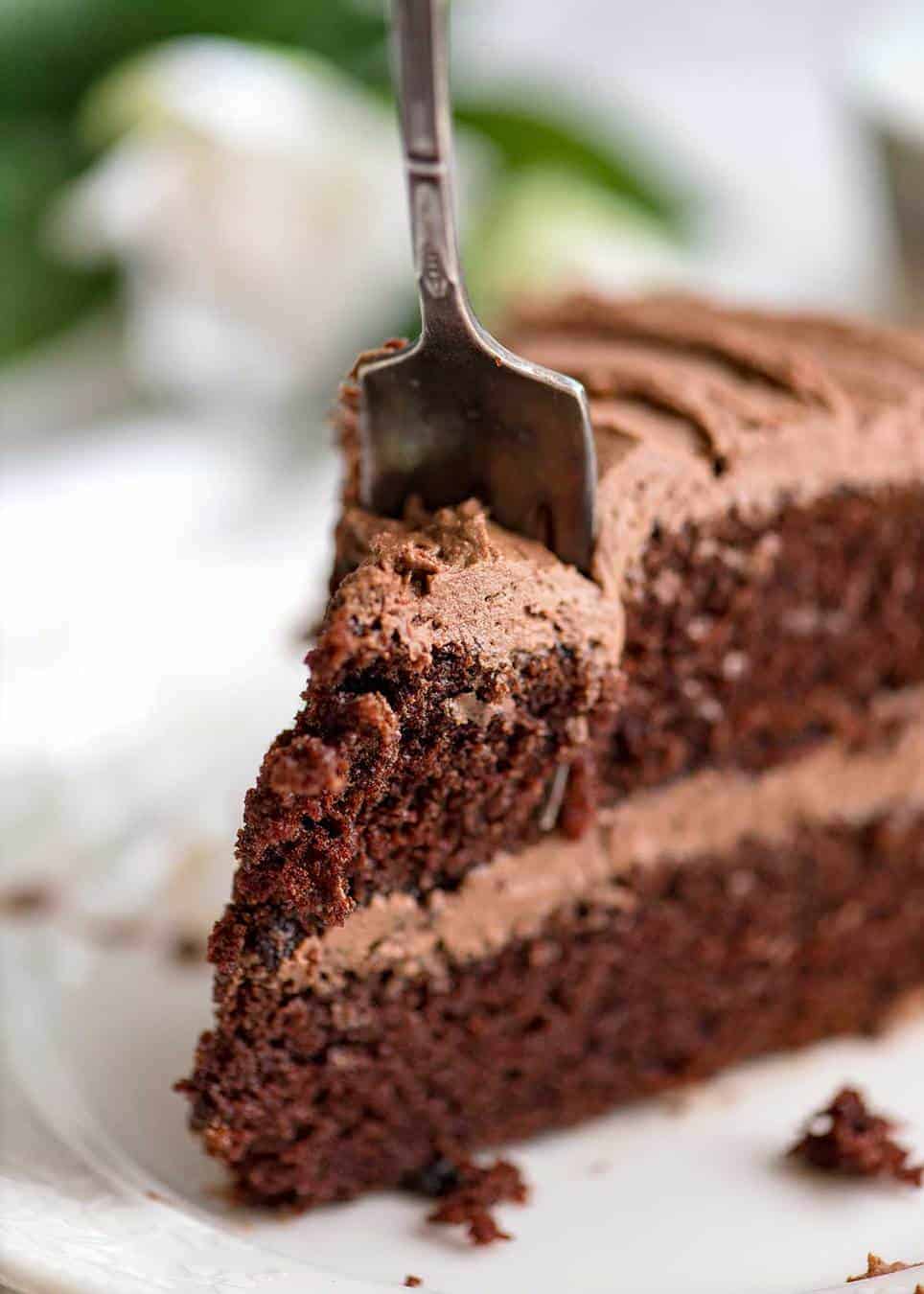 Close up photo of a slice of Chocolate Cake with Buttercream Chocolate Frosting on a white dessert plate with a fork partially cutting into it.