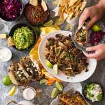 Overhead photo of Mexican Fiesta taco dinner party, with pork carnitas, tacos, grilled corn, guacamole, salsa, pickled red cabbage, tortilla chips and taco sides.
