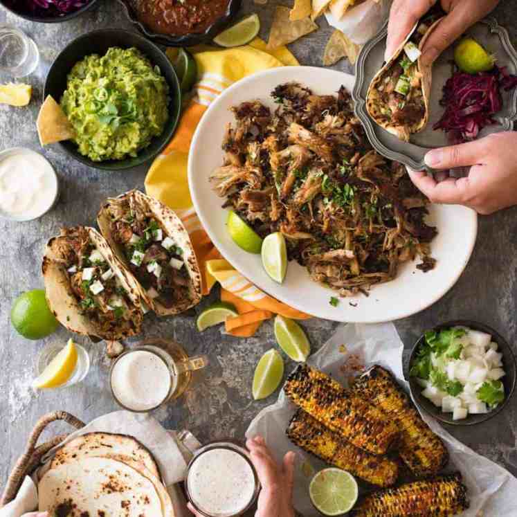 Overhead photo of Mexican Fiesta taco dinner party, with pork carnitas, tacos, grilled corn, guacamole, salsa, pickled red cabbage, tortilla chips and taco sides.