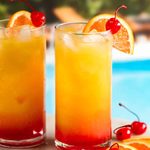 Zwei Tequila Sunrise Cocktails an einem Pool, garniert mit Orangenscheibe und Maraschino-Kirschen trinkfertig